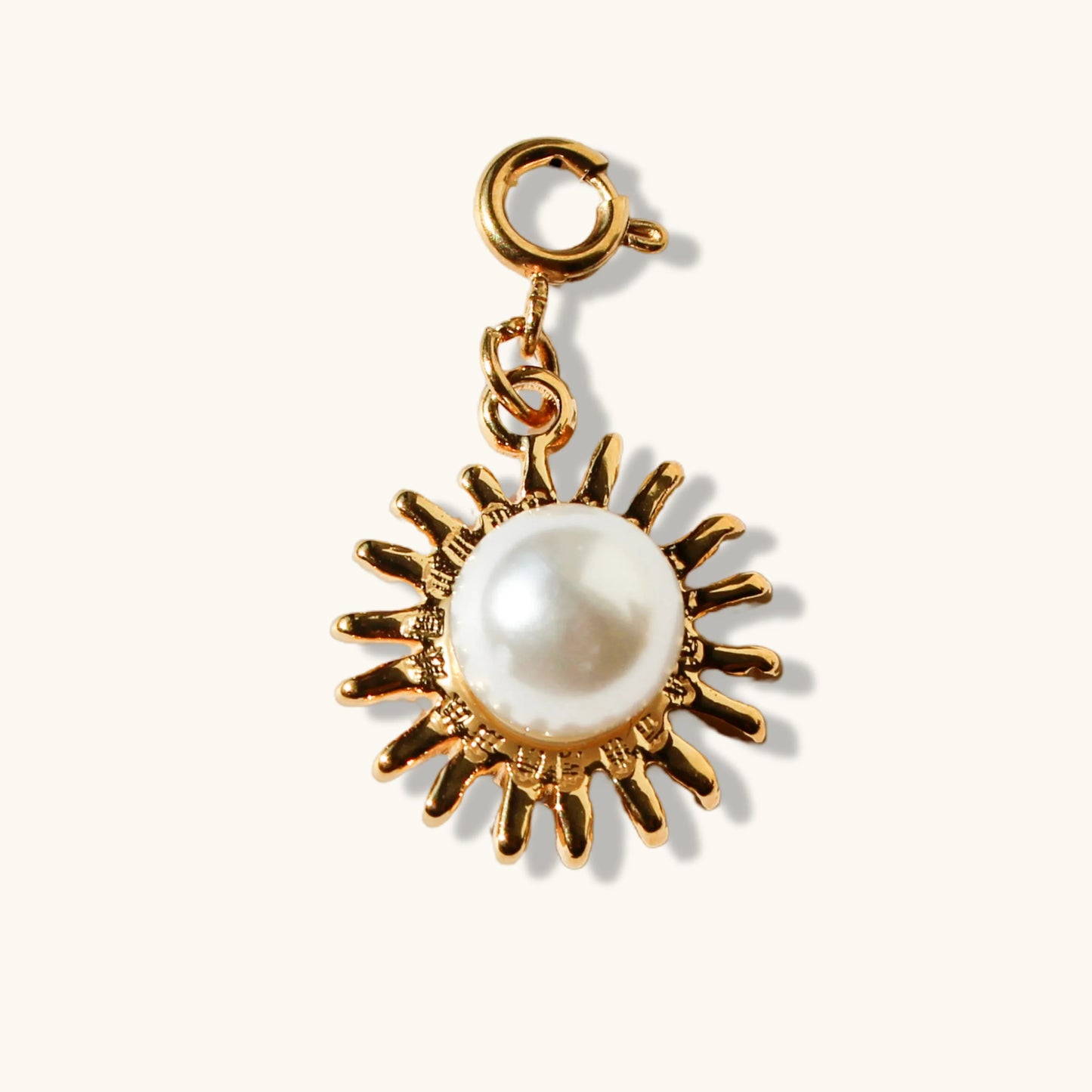 Gold sun charm with white pearl center on cream background, jewelry accessory