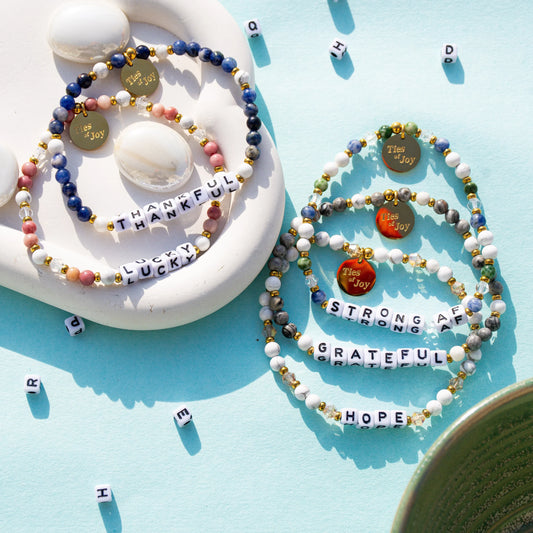 Colorful beaded bracelets with uplifting words like thankful, lucky, strong, and grateful on a blue background.