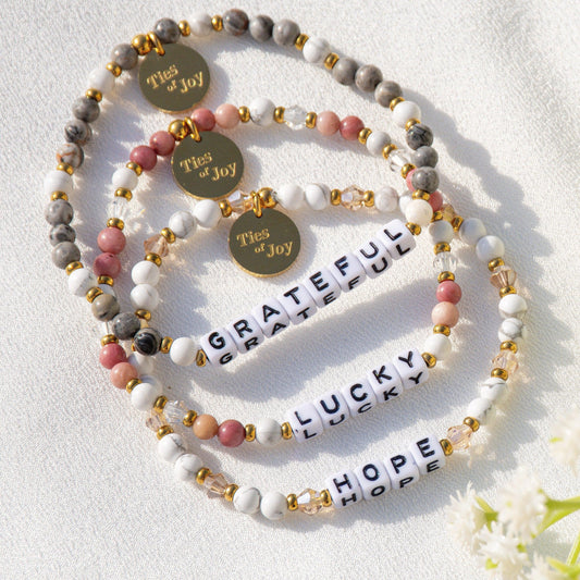 Ties of Joy beaded bracelets with 'GRATEFUL,' 'LUCKY,' and 'HOPE' letter beads on fabric