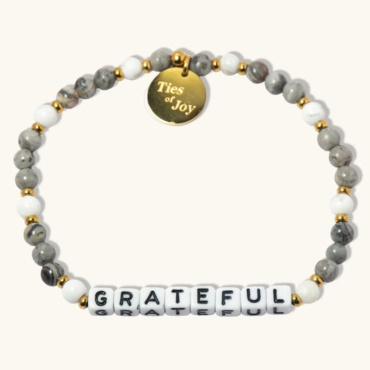 Gray and white beaded bracelet with gold charm reading 'Ties of Joy' and 'GRATEFUL' letter beads.
