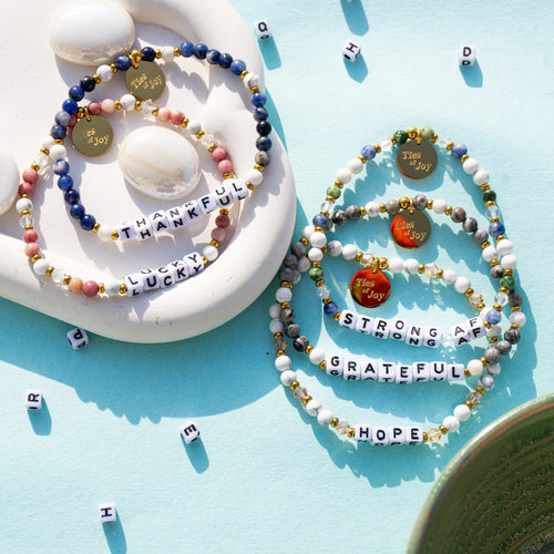 Colorful beaded bracelets with uplifting words like thankful, lucky, strong, and grateful on a blue background.
